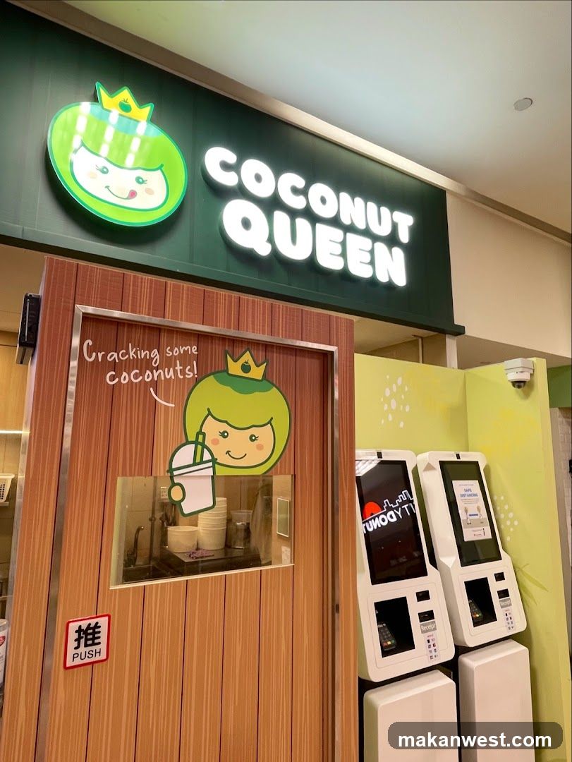 The exterior of a Coconut Queen shop with a prominent sign above. The sign features a cartoon coconut character wearing a crown and the text "COCONUT QUEEN" in illuminated letters. Below the sign, a wooden-textured door has a window revealing a preparation area with cups and a sink. A "PUSH" sign is visible on the door. To the right, two self-ordering kiosks are present.