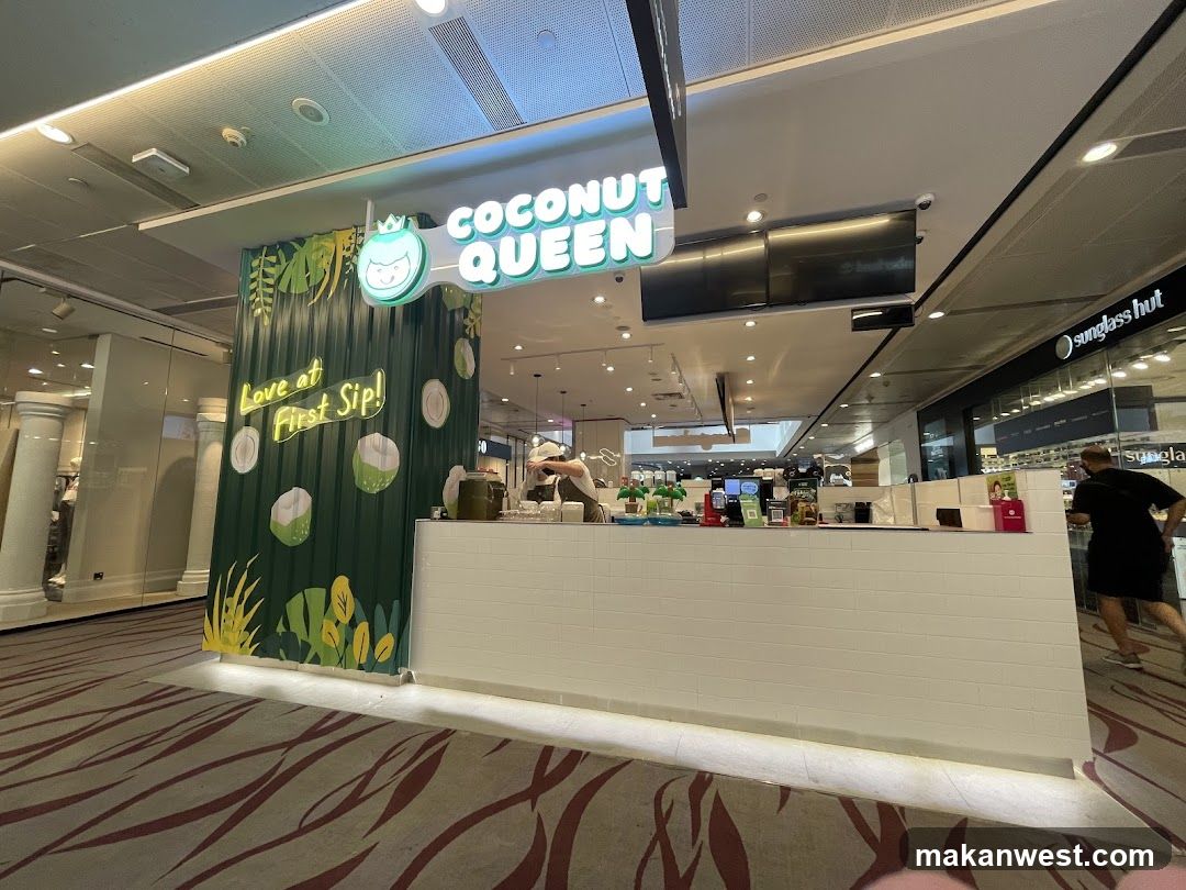 The exterior of a Coconut Queen shop with a white tiled counter and a green corrugated metal wall decorated with illustrations of coconuts and leaves. The shop's name is displayed in neon lights above the counter. A staff member is visible behind the counter preparing a drink.