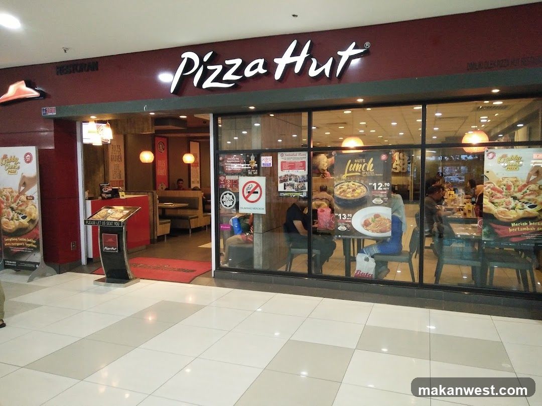 The exterior of a Pizza Hut restaurant with large glass windows. The Pizza Hut logo is prominently displayed above the entrance. Inside, tables and booths are visible, with some customers seated. Promotional posters for pizzas and lunch specials are displayed on the windows and outside the entrance.