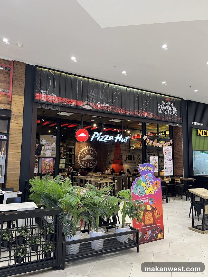 The exterior of a Pizza Hut restaurant with its signature red and white logo and signage visible. The restaurant is located inside a mall, with other businesses visible next to it. There are tables and chairs inside the restaurant, and people are dining. A promotional standee featuring Pokemon characters and pizzas is placed outside the entrance.