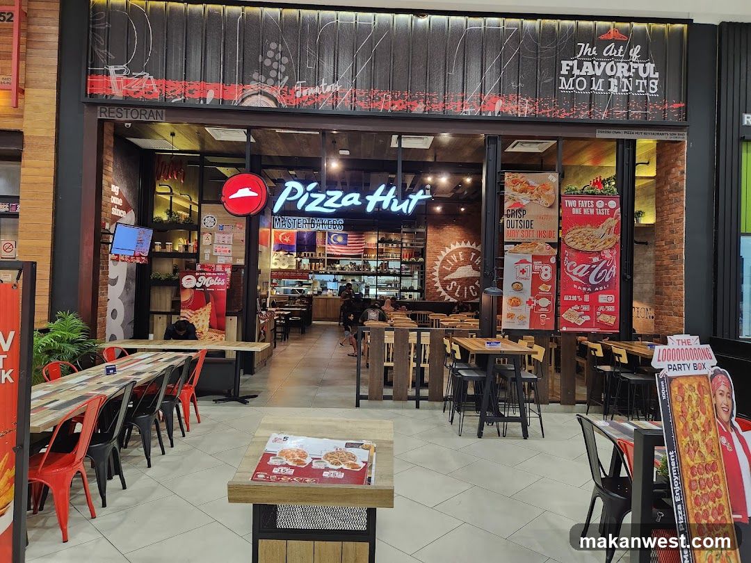 The exterior of a Pizza Hut restaurant with tables and chairs visible both inside and outside. Promotional posters are displayed on the windows and a cardboard cutout of a pizza is near the entrance.