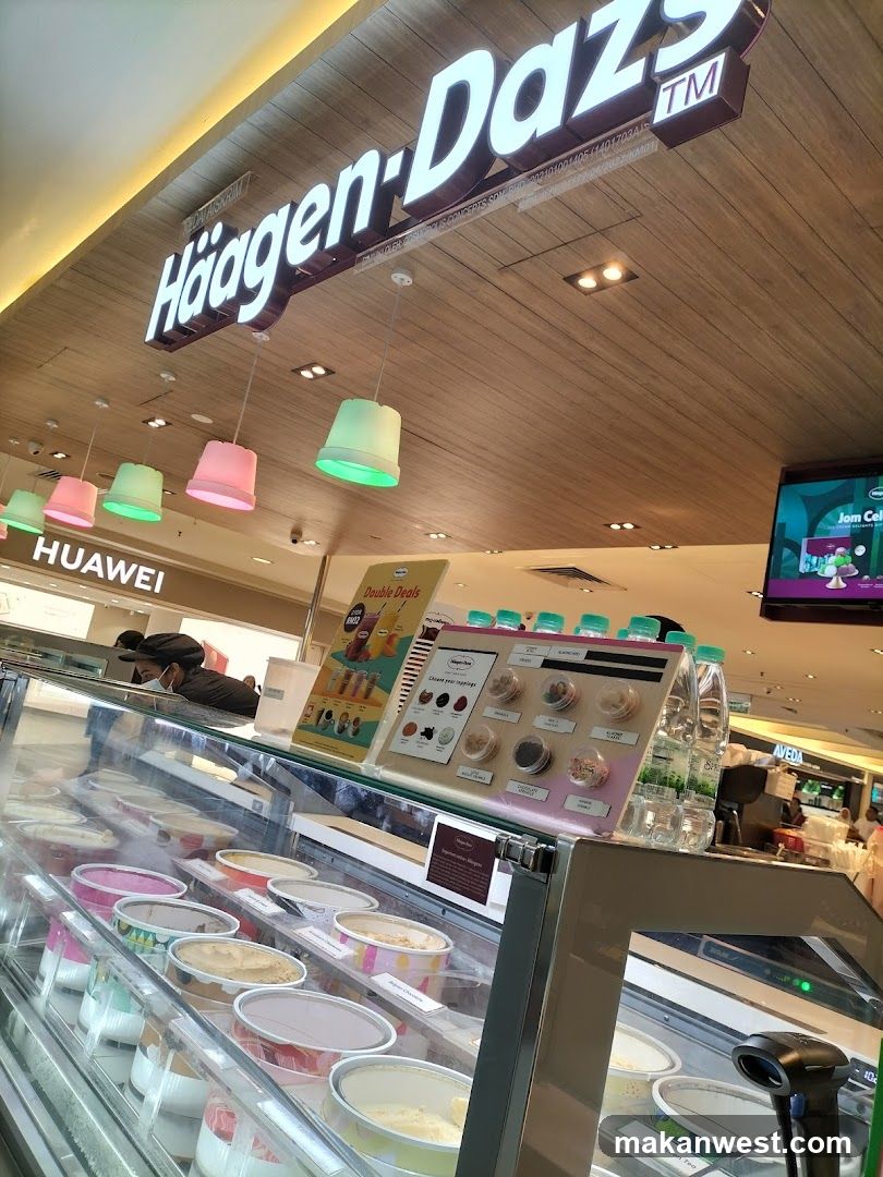 A Haagen-Dazs ice cream shop with a display case full of ice cream tubs and a prominent Haagen-Dazs sign above. There are colorful hanging lights and a menu board displaying toppings.