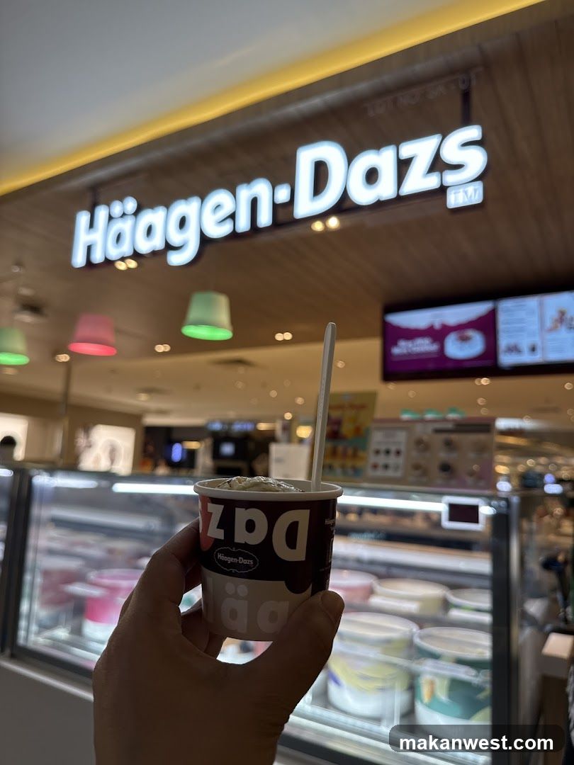 A hand holds a small cup of ice cream in front of a Haagen-Dazs store. The store's illuminated sign is visible above. Inside, ice cream tubs are displayed in a refrigerated case.