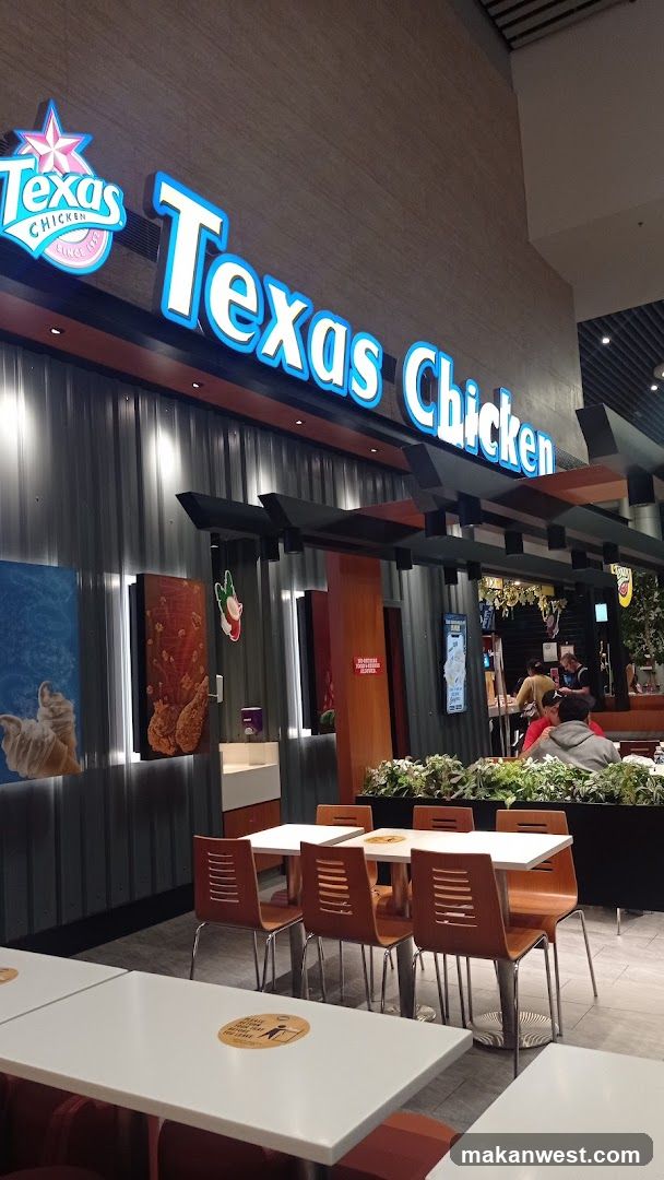 A Texas Chicken restaurant storefront with illuminated signage displaying the brand name and logo. The facade features vertical metal paneling, decorative artwork of chicken pieces, and a seating area outside with white tables and orange chairs. A few customers are visible near the entrance.