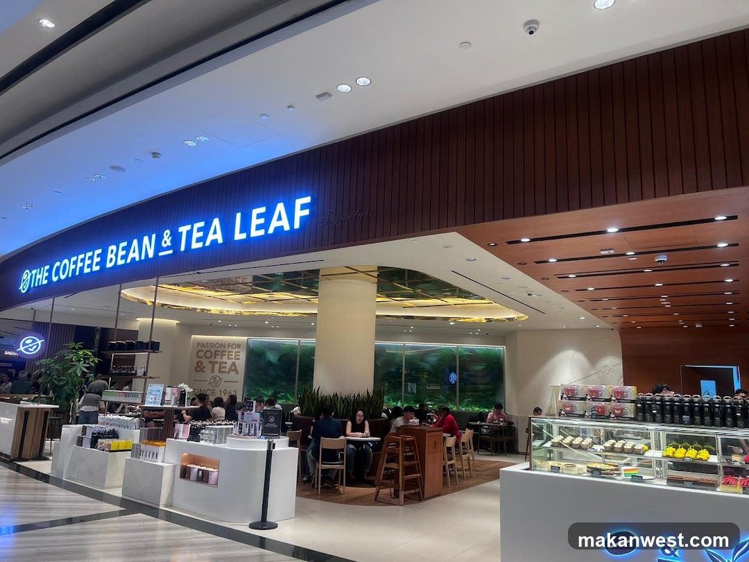 A modern coffee shop entrance with illuminated signage reading 'THE COFFEE BEAN & TEA LEAF'. The storefront features a sleek design with wooden paneling and glass windows, showcasing an inviting interior where customers are seated at tables enjoying their beverages. A display case outside highlights various baked goods and products available for purchase.