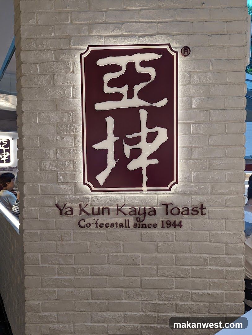A close-up of a white brick wall featuring a large illuminated sign with Chinese characters '亞坤' and the English text 'Ya Kun Kaya Toast Co'teestall since 1944'. The sign is mounted on the exterior facade, indicating it is part of a shop front.