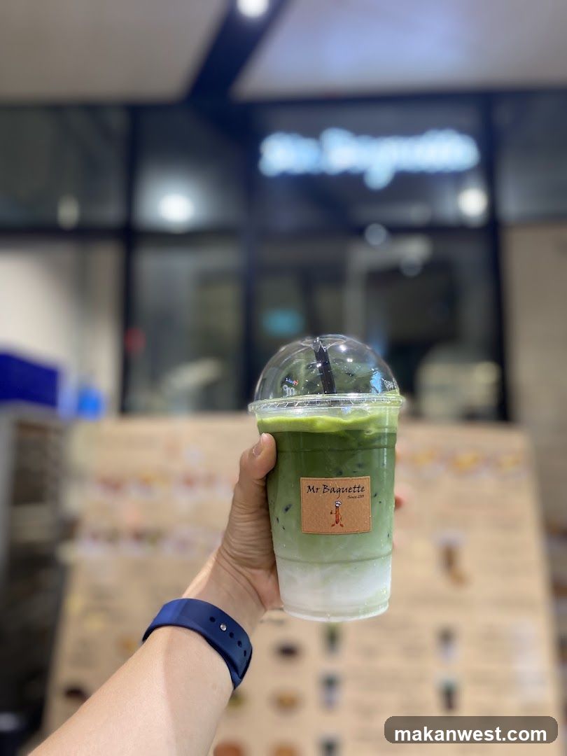 A hand holding a clear plastic cup with green matcha drink and white foam at the bottom, featuring a label reading 'Mr Biscotti'. Background shows blurred interior of a shop with menu boards on display.