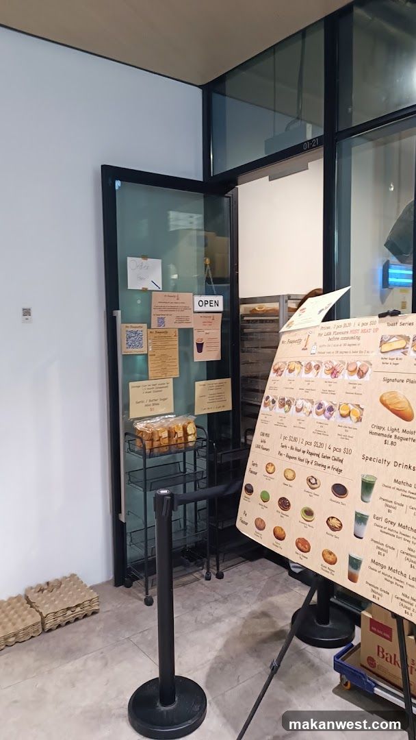 Interior shot of a bakery showing a glass door entrance with 'OPEN' sign and notices, a rack of packaged breads, cardboard egg trays on the floor, and a large menu board displaying pastries, drinks, and prices.