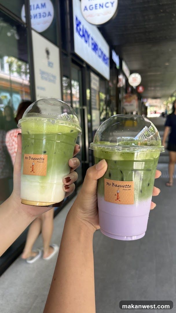 Two hands holding plastic cups with layered green (matcha) and purple (lavender) drinks, each labeled 'Mr Baguette'. Background shows storefront signage including 'AGENCY' and glass windows reflecting street activity.