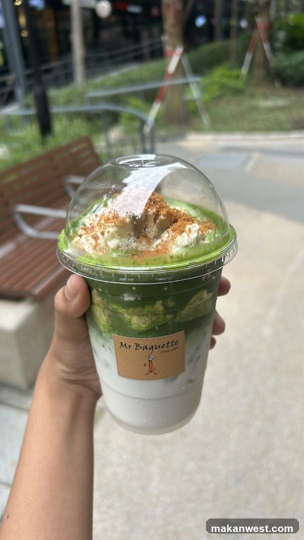 A hand holding a clear plastic cup filled with layers of white milk and green matcha, topped with whipped cream and crumbled brown topping. The cup has a dome lid and a label reading 'Mr Baguette'.