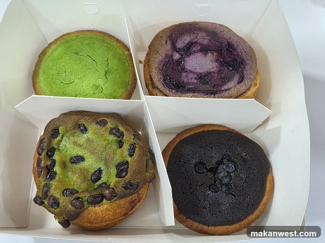 A white cardboard box containing four round pastries arranged in two rows of two. Top left is green (matcha?), top right is purple (blueberry or ube), bottom left is green with dark chocolate chips, and bottom right is dark brown/black (chocolate). All appear to be baked goods with slightly cracked surfaces.