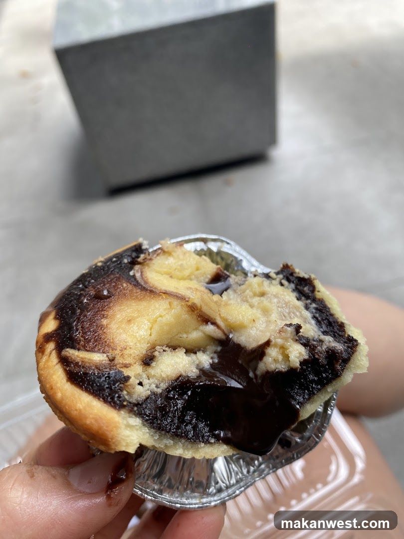 A hand holds a partially eaten pastry with chocolate swirls and cream filling, served in a foil cup.