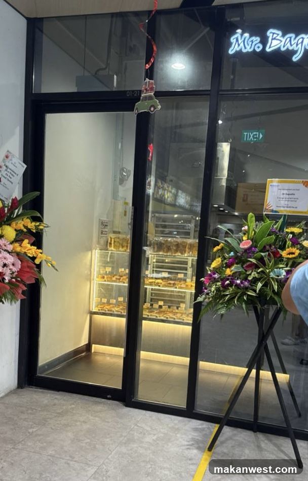 Exterior view of a bakery storefront with glass doors and windows displaying baked goods inside. Neon sign reading 'Mr. Bag' is visible above the entrance. Two floral arrangements flank the doorway, one on each side. A small decorative car hangs from the top center of the door frame. Inside, shelves hold trays of pastries under warm lighting. A yellow poster is affixed to the right window pane.