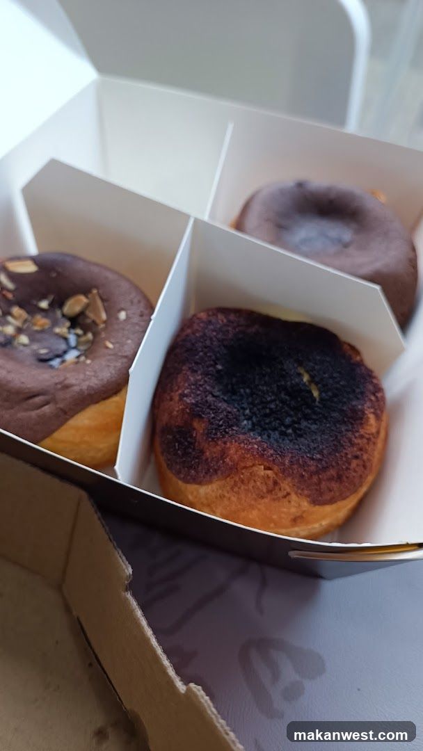A white cardboard box containing four pastries, each in its own compartment. The pastries appear to be donuts or similar baked goods with different toppings and glazes.