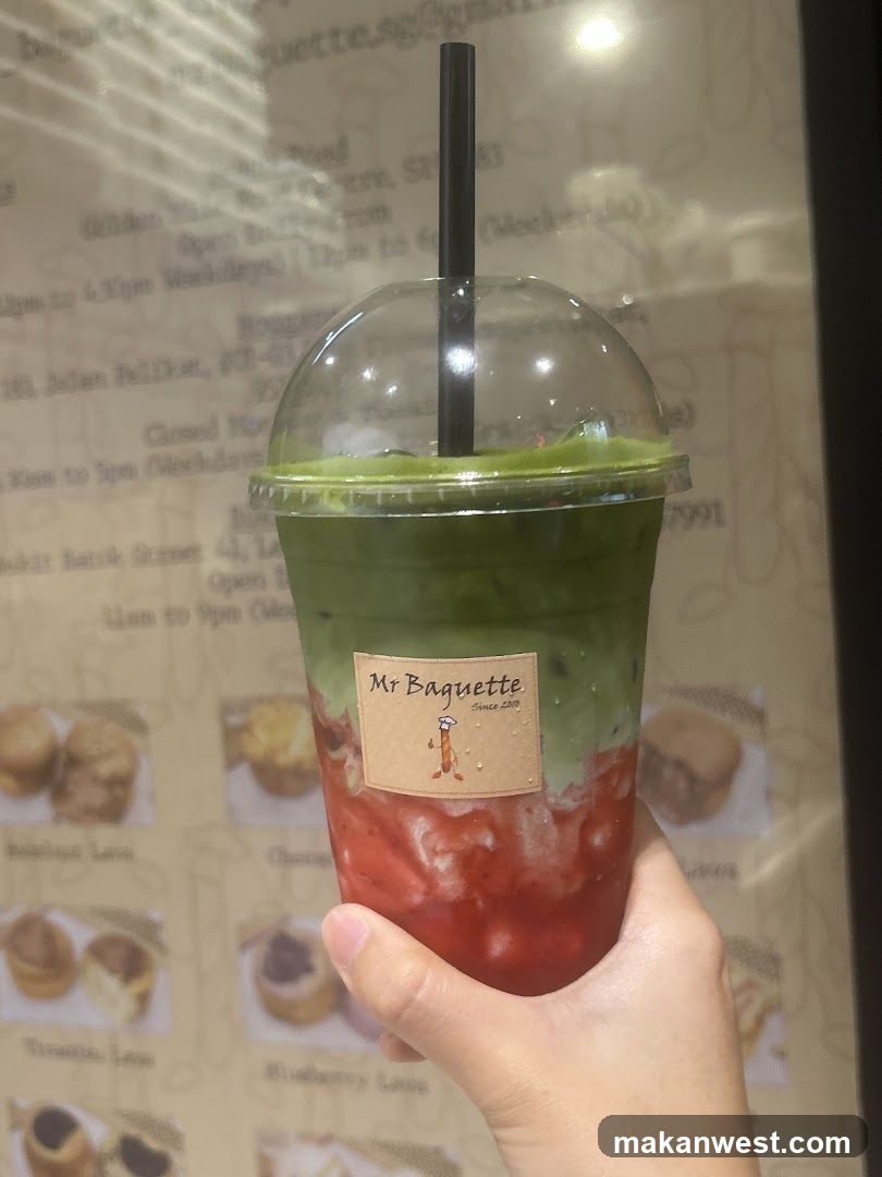 A hand holds a layered iced drink with green matcha on top and red strawberry base in a clear plastic cup with dome lid and black straw. The cup has a label reading 'Mr Baguette Since 2019' with a cartoon character. Background shows a menu board listing items like 'Breakfast Loaf', 'Choco Loaf', 'Blueberry Loaf', 'Tiramisu Loaf', and addresses including Golden Mile Road, Jalan Jelikat, and Bukit Bintang Street.