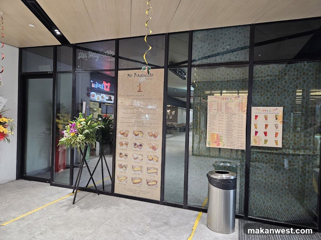 A modern glass-fronted bakery entrance with a large menu board on the door, flower arrangements at the entrance, and a trash bin outside.