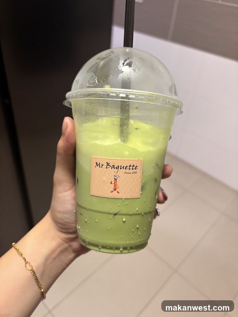 A hand holding a clear plastic cup filled with green matcha drink, featuring ice cubes and condensation on the outside. The cup has a dome lid with a black straw inserted. A label reads 'Mr Baguette' with a logo of a baguette character.