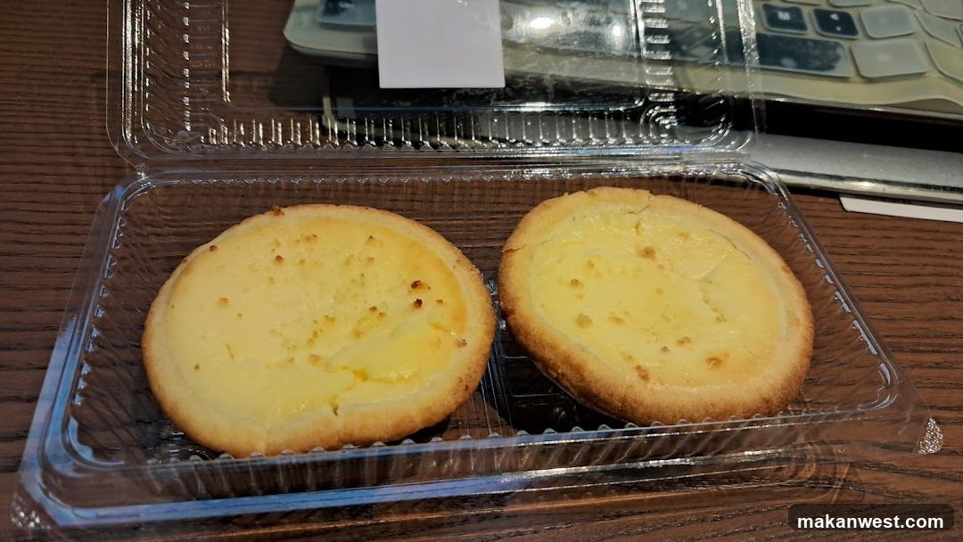 Two golden-brown custard tarts with smooth yellow filling are placed side by side inside a clear plastic takeout container. The container rests on a wooden table surface, with part of a keyboard visible in the background.