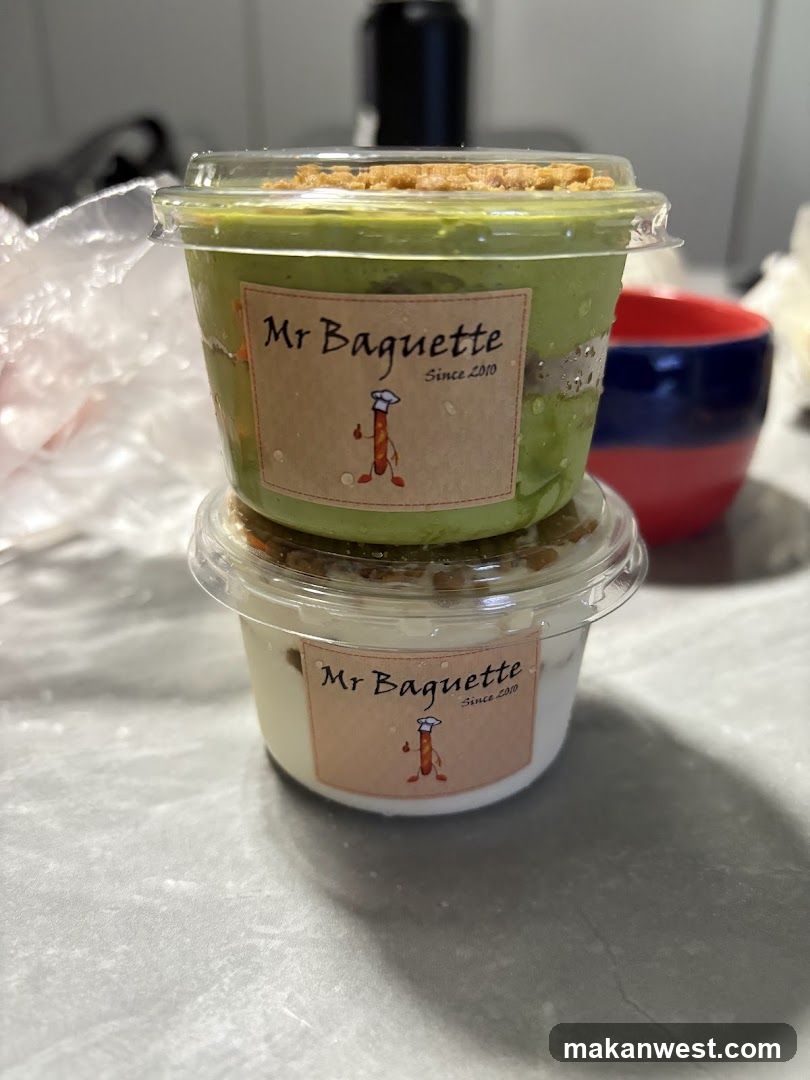 Two clear plastic dessert cups stacked on a countertop, each labeled with 'Mr Baguette Since 2019' and a cartoon baguette character. The top cup contains green pudding or mousse topped with crumbled nuts; the bottom cup holds white cream or yogurt with similar topping. Background includes blurred kitchen items.