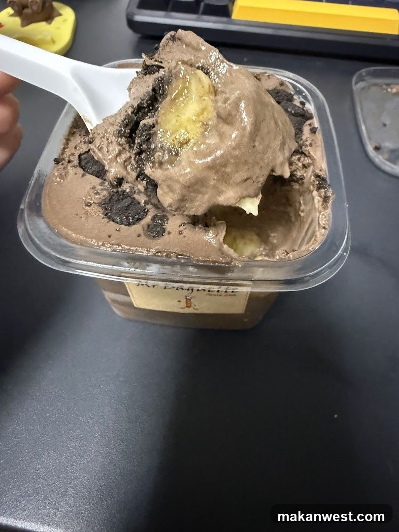 A close-up of a plastic container filled with chocolate ice cream, topped with banana slices and cookie crumbles. A white spoon scoops up a portion of the dessert. The container has a label that reads 'MY PAGHULLIS'. In the background, there is a keyboard and some other items on a dark surface.
