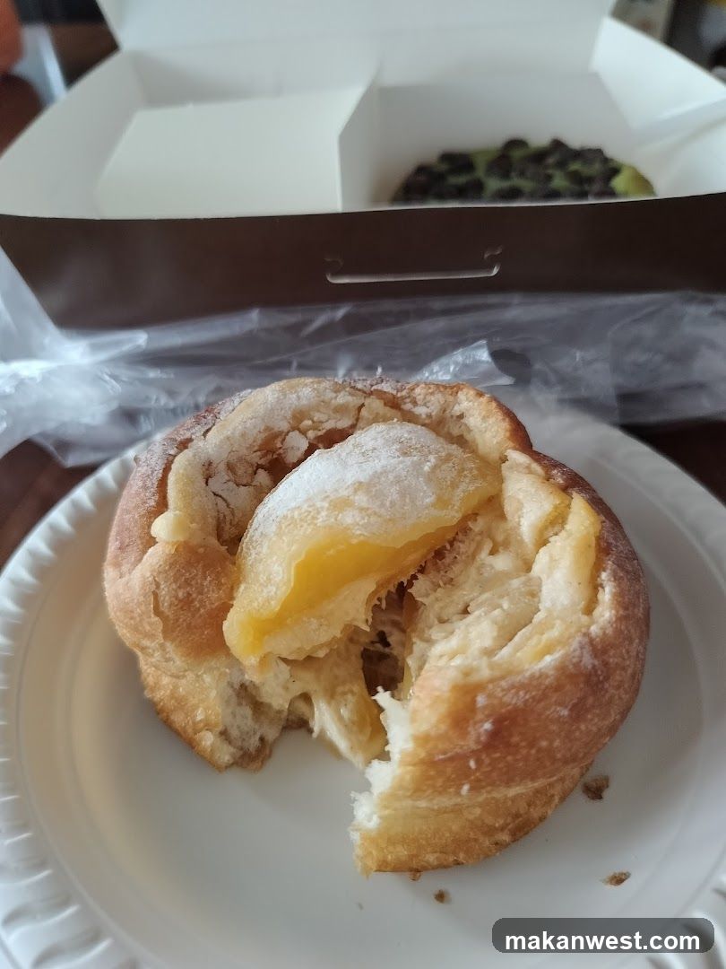 A close-up of a partially eaten pastry on a white plate, featuring a golden-brown crust and visible lemon filling. In the background, there is an open box containing another pastry.