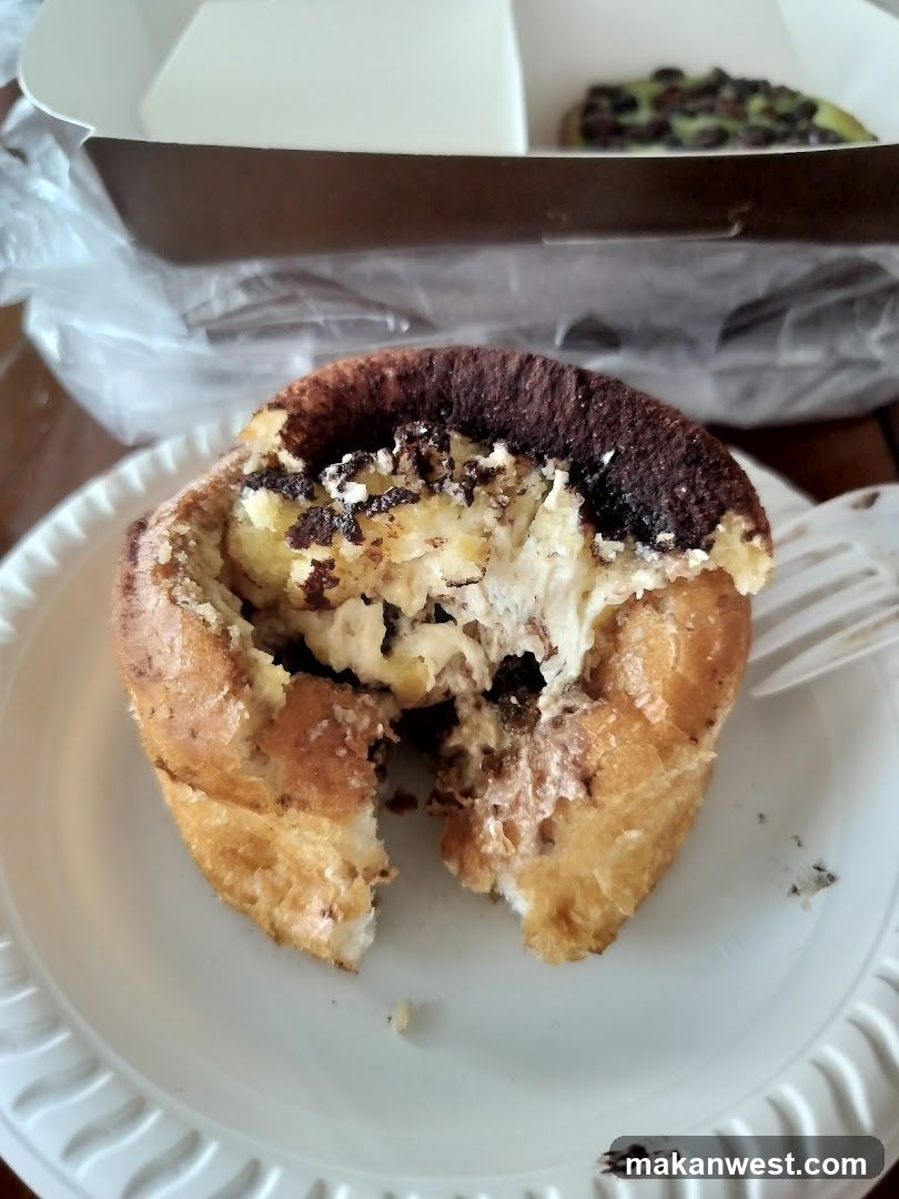A partially eaten pastry with a golden-brown crust and creamy filling, served on a white paper plate. In the background, there is a plastic bag containing another pastry.