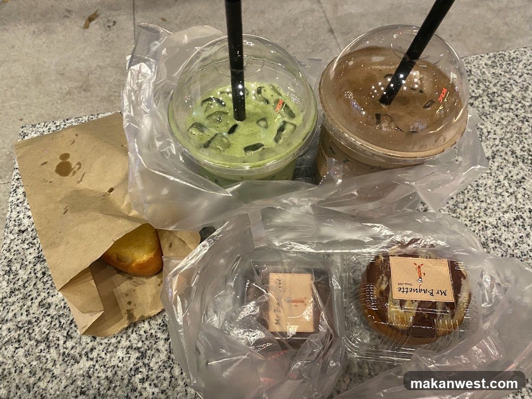 A close-up of a takeout meal featuring two iced drinks (green matcha and brown iced coffee) in plastic cups with straws, wrapped in clear plastic bags. Two packaged pastries are visible—one labeled 'Mr Paquette'—and a partially opened paper bag containing what appears to be a bread roll or pastry. The items rest on a speckled countertop.
