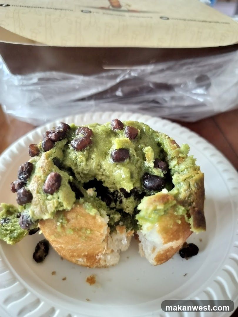 A partially eaten bun filled with green matcha paste and topped with dark brown beans, served on a white disposable plate. A cardboard box is visible in the background.