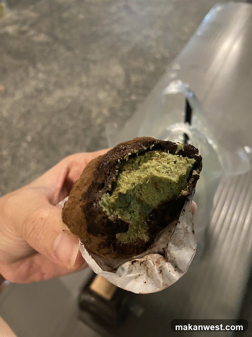 A hand holds a partially eaten pastry with green matcha filling and dark brown exterior, wrapped in white paper.