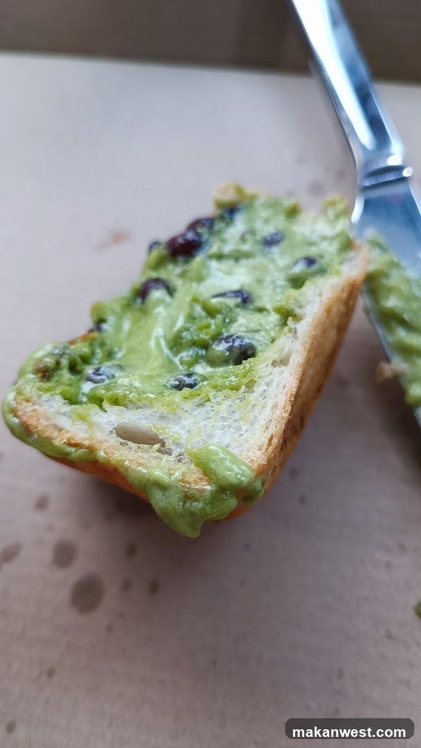 A close-up of a slice of bread topped with mashed avocado and blueberries, spread unevenly on the surface. A knife is visible in the background.