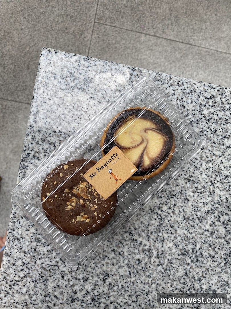 A clear plastic container holding two pastries on a speckled granite surface. One pastry is chocolate with chopped nuts, the other has a swirl of light and dark filling. A small card labeled 'Mr Baquette' sits between them.
