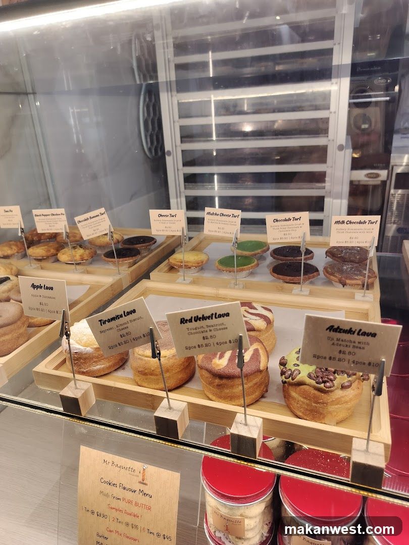 A glass display case filled with various pastries and cakes, each labeled with a small sign indicating the name and price. The items include Apple Lava, Tiramisu Lava, Red Velvet Lava, Adzuki Lava, Cheese Tart, Matcha Cheese Tart, Chocolate Tart, and Milk Chocolate Tart. In the background, there is an oven and other kitchen equipment visible through the glass.