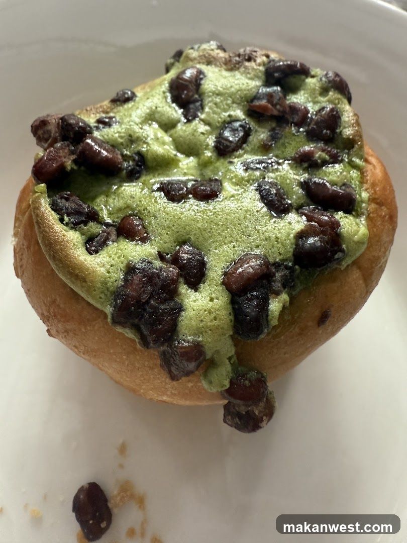 A close-up of a golden-brown bun topped with green matcha paste and dark bean pieces, served on a white plate.