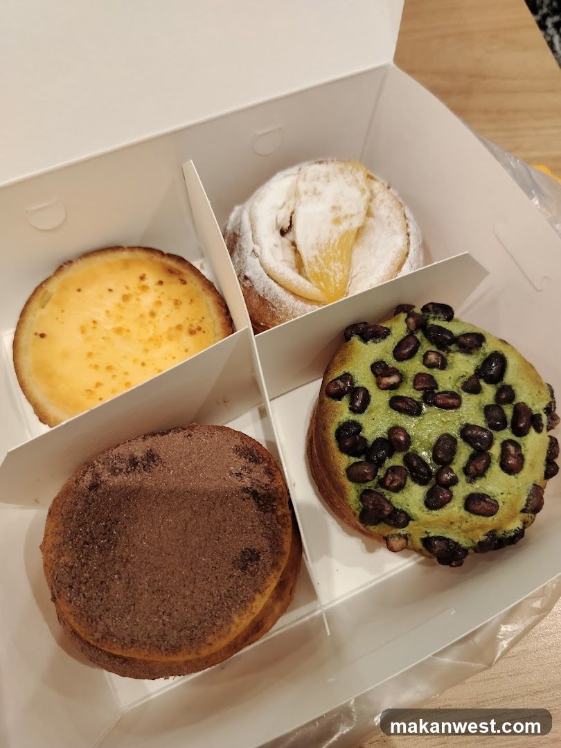 A white box containing four different pastries: a custard tart, a powdered donut with lemon filling, a chocolate-dusted pastry, and a green matcha-flavored pastry topped with dark beans.
