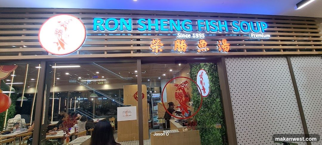 The image shows the exterior of a shop named 'RON SHENG FISH SOUP' with Chinese characters below it. The sign is illuminated and mounted on a wooden slat facade. There are glass windows showing the interior, where tables and chairs are visible. A red lantern hangs to the left side. Inside, there's a counter area with staff members working behind it. Decorative elements include greenery panels and circular signs featuring fish imagery.