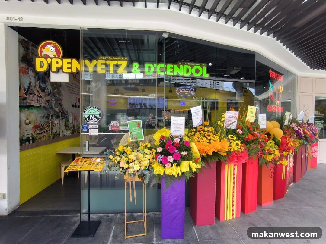 A vibrant storefront featuring a glass facade with neon signage reading 'D'PENYEZ & D'CENDOL'. The entrance is adorned with colorful floral arrangements in red, yellow, and purple vases. Inside, the interior displays bright yellow walls and shelves stocked with products. A small table with promotional materials stands near the entrance.