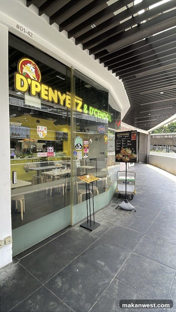 The image shows the exterior of a shop named 'D'PENYEZ & D'CENDORI', featuring large glass windows and doors with bright yellow signage. Inside, tables and chairs are visible along with kitchen equipment. A menu board stands outside near the entrance.