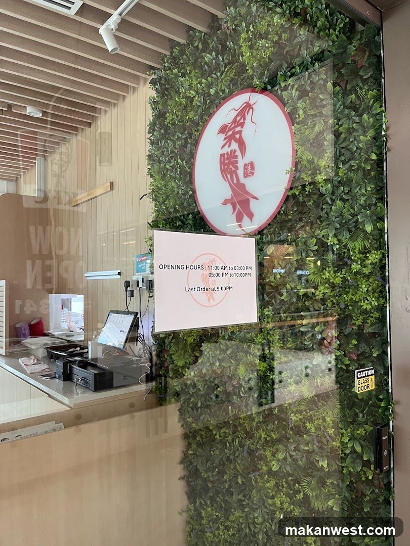 A modern restaurant entrance featuring a glass door and window framed by a lush green vertical garden wall. A circular red logo with stylized Chinese characters is mounted above the door. An open hours sign indicates service from 11 AM to 2 PM and 5 PM to 10 PM, with last order at 8 PM. Inside, a counter with cash register equipment is visible through the glass.