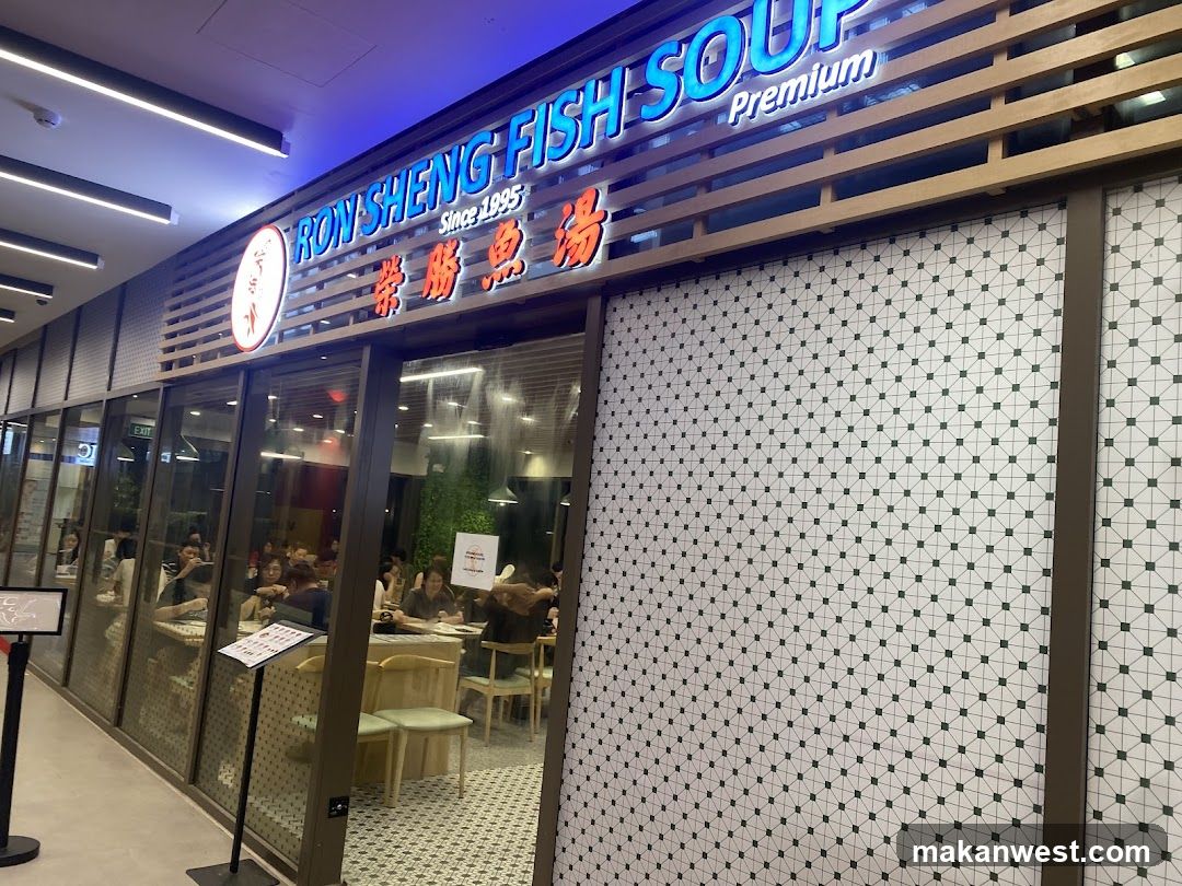 The image shows the exterior of a restaurant named 'Ron Sheng Fish Soup Premium'. The sign is illuminated with blue and red lights, featuring both English and Chinese characters. Inside, patrons are seated at tables, enjoying their meals.