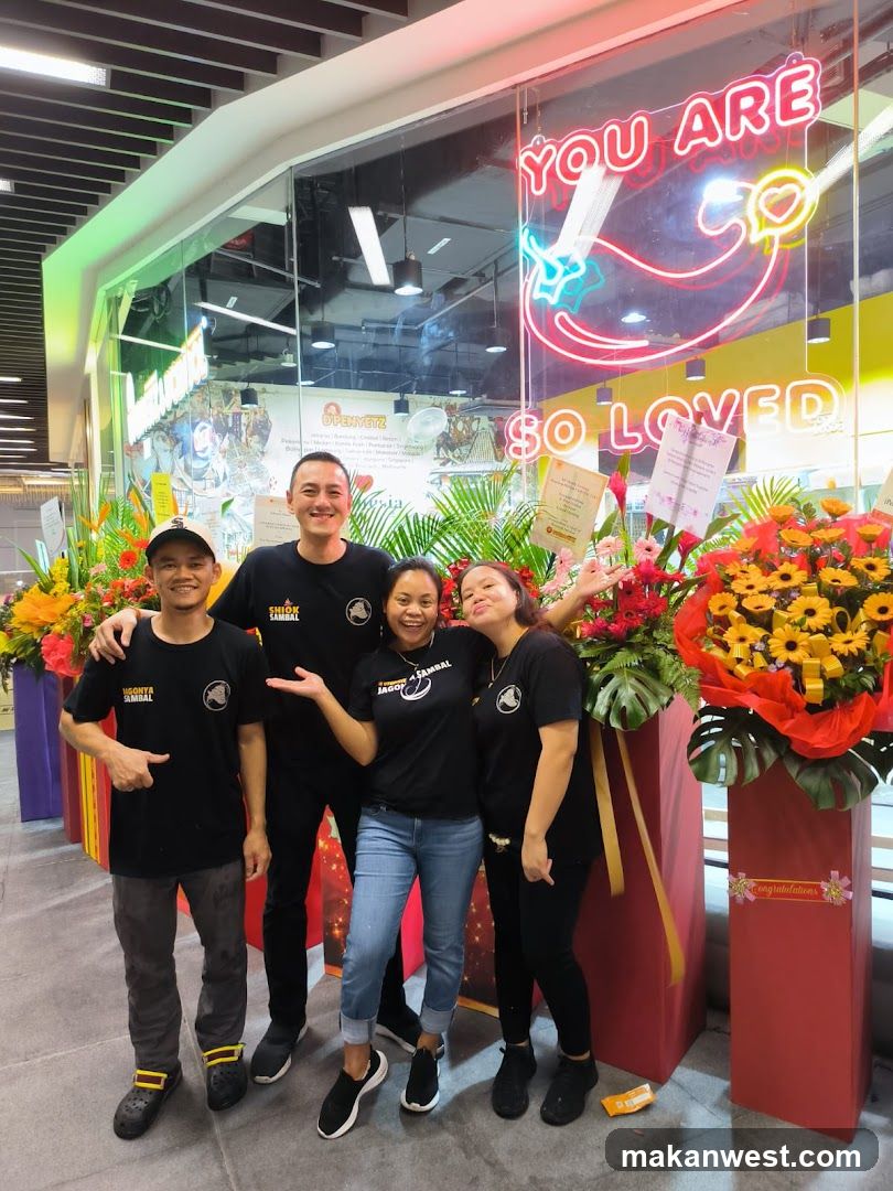 Group of five staff members posing in front of a brightly lit storefront with neon signage reading 'YOU ARE SO LOVED' and a heart graphic. Flower arrangements flank the entrance. Staff wear matching black t-shirts with store logo.