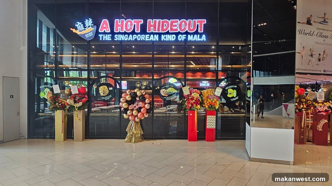 The storefront features a large glass facade with illuminated signage reading 'A HOT HIDEOUT' and the tagline 'THE SINGAPOREAN KIND OF MALA'. Several floral arrangements and balloon displays are placed at the entrance, indicating a recent opening or celebration. Inside, warm lighting illuminates the dining area.