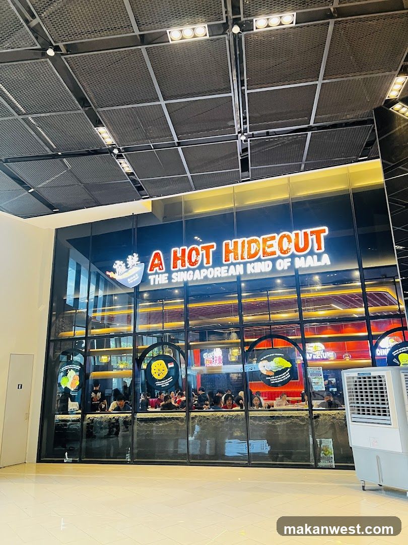 The storefront features a large glass facade with the illuminated sign 'A HOT HIDEOUT' and subtitle 'THE SINGAPOREAN KIND OF MALA'. Inside, customers are seated at tables under warm lighting.
