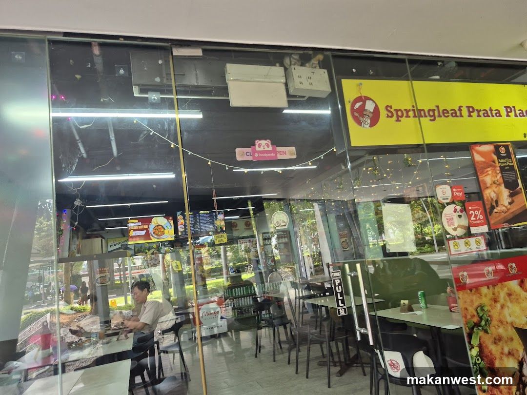 Photo of physical exterior entrance/storefront facade with visible signage 'Springleaf Prata Place' on yellow background, glass doors, interior seating area, and promotional posters.