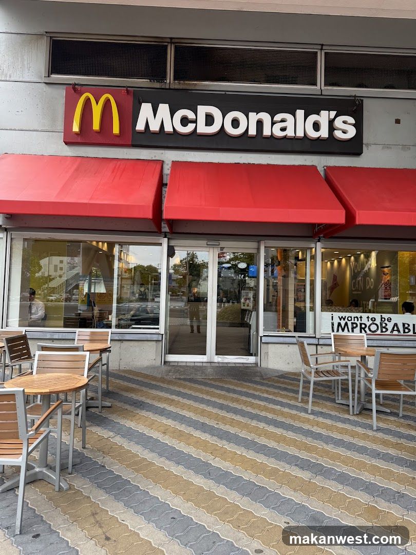 Photo of physical exterior entrance/storefront facade with visible shop signage ON the building. Red awnings, glass doors and windows, outdoor seating area on striped pavement.
