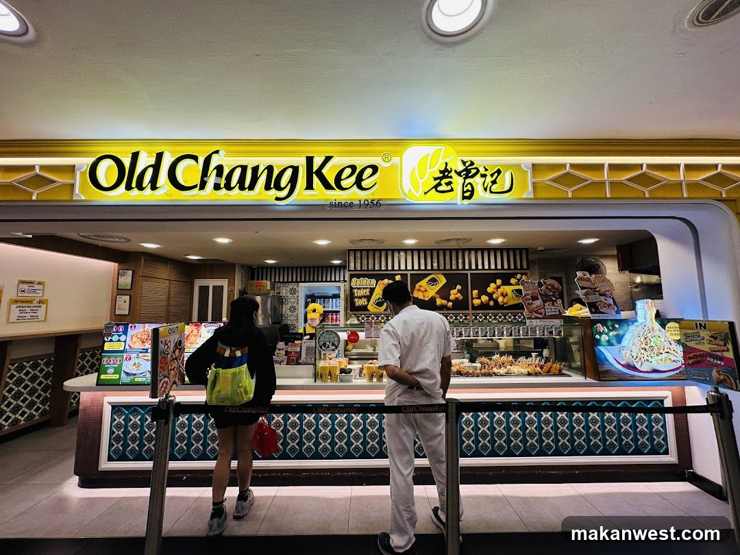 A brightly lit shop front with a yellow and white facade featuring the 'Old Chang Kee' logo in English and Chinese characters. The entrance is open, revealing an interior counter where staff are preparing food. Two customers stand at the counter, one wearing a black jacket and carrying a green bag, while another wears a white uniform. Inside, shelves display various products including packaged snacks and beverages. A digital screen shows promotional content on the right side.