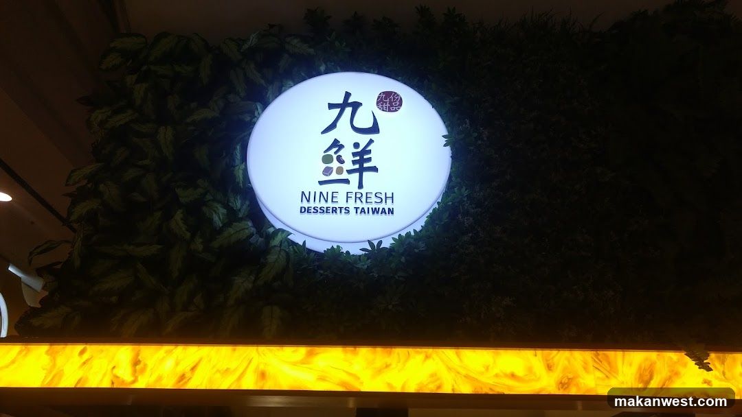 A brightly lit circular sign featuring Chinese characters '九鮮' and English text 'NINE FRESH DESSERTS TAIWAN', surrounded by green foliage against a dark background, with yellow illuminated panel below.