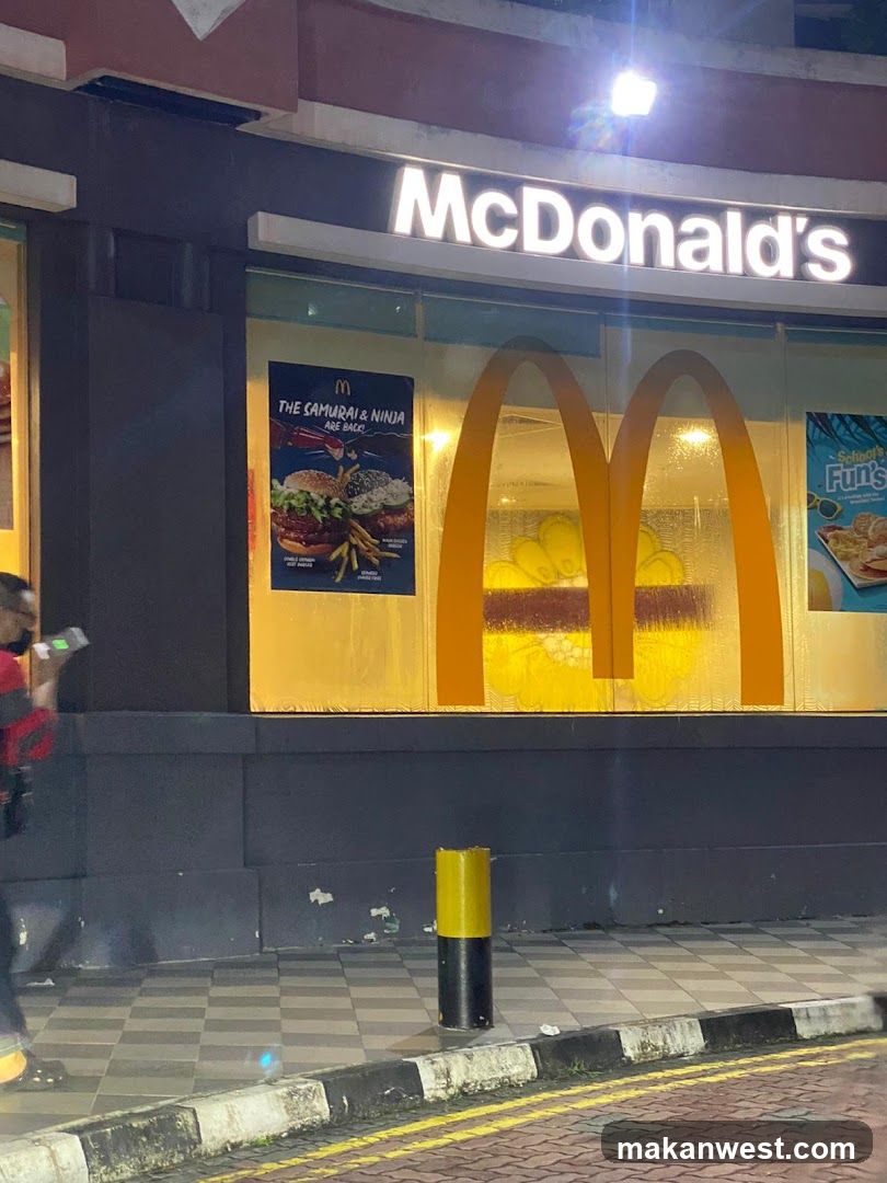 A nighttime photo of a McDonald's storefront featuring illuminated signage and large golden arches in the window. A poster advertises 'The Samurai & Ninja' burger combo, while another promotes 'Fun!'. The sidewalk is checkered with yellow-black curb markings visible.