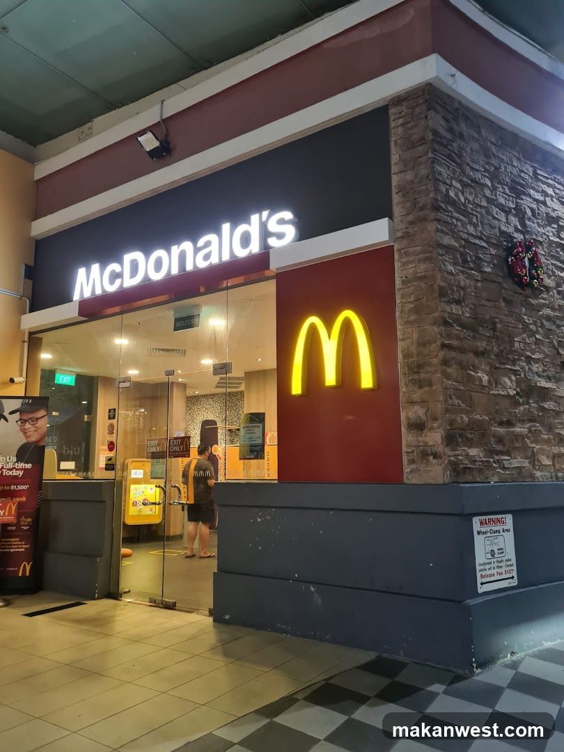 Photo of the physical exterior entrance and storefront facade of a McDonald's restaurant. The building features a prominent 'McDonald's' sign with illuminated lettering above glass doors, alongside a large glowing yellow 'M' logo on a red panel. A stone-textured wall section is visible to the right, adorned with a small wreath. Inside, through the glass, a customer in uniform stands near an ordering kiosk. To the left, a promotional poster for 'PayNow' is displayed outside.