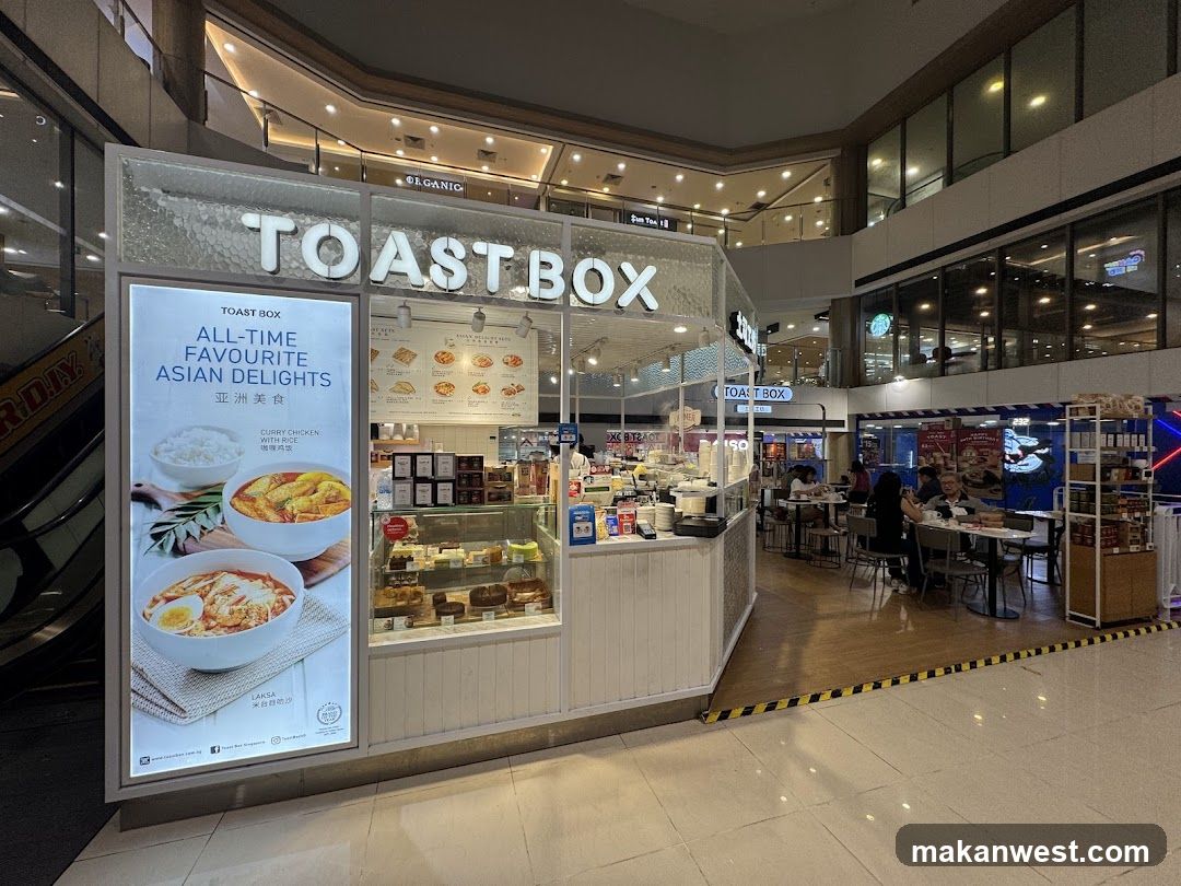 A brightly lit shop front of TOAST BOX, featuring a large illuminated sign with 'TOAST BOX' lettering above the entrance. A vertical poster on the left displays Asian dishes like curry chicken and egg rice bowls. Inside, glass display cases show baked goods and pastries. The interior is visible through open doors, showing customers seated at tables in the background. The floor is tiled, and an escalator is partially visible to the left.