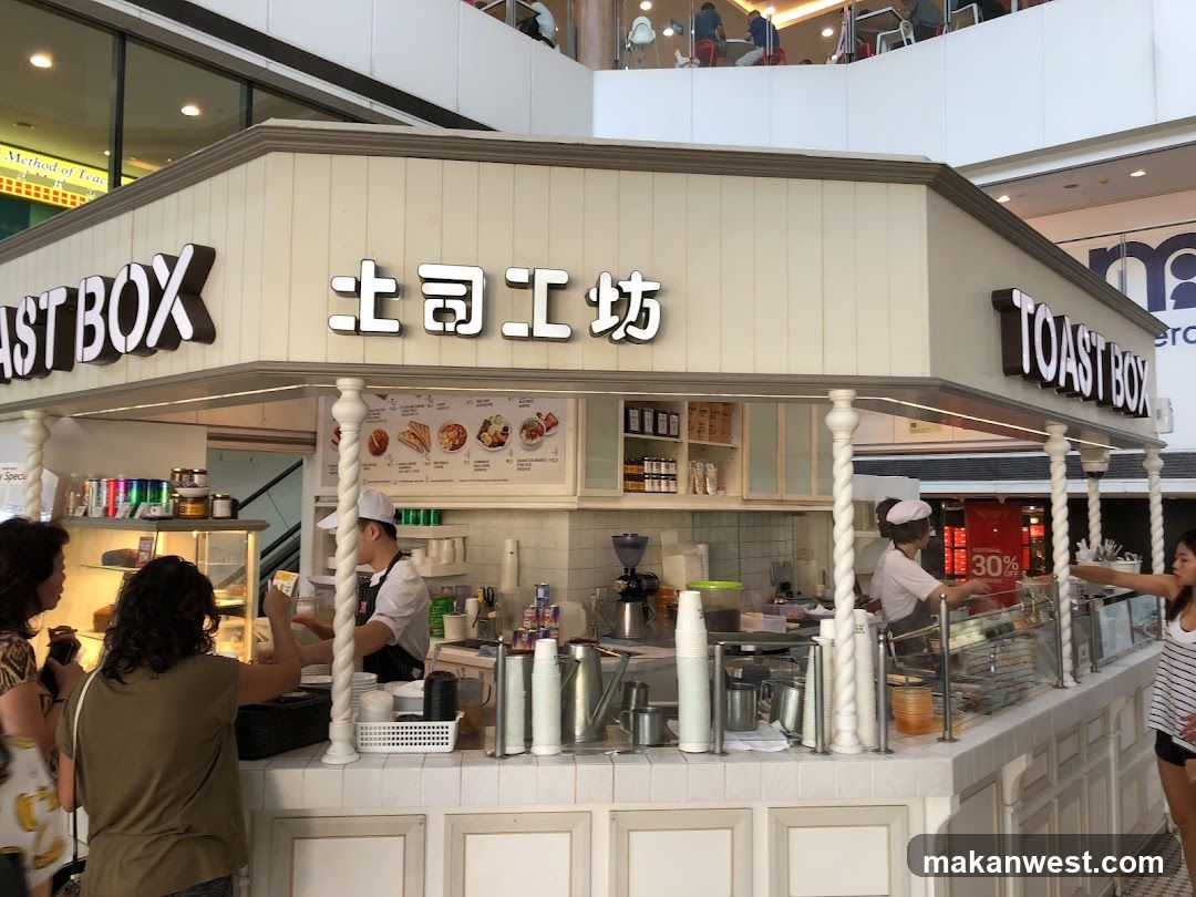 A food stall with 'TOAST BOX' and Chinese characters on the facade, featuring staff preparing food behind a counter with various kitchen equipment and ingredients visible.